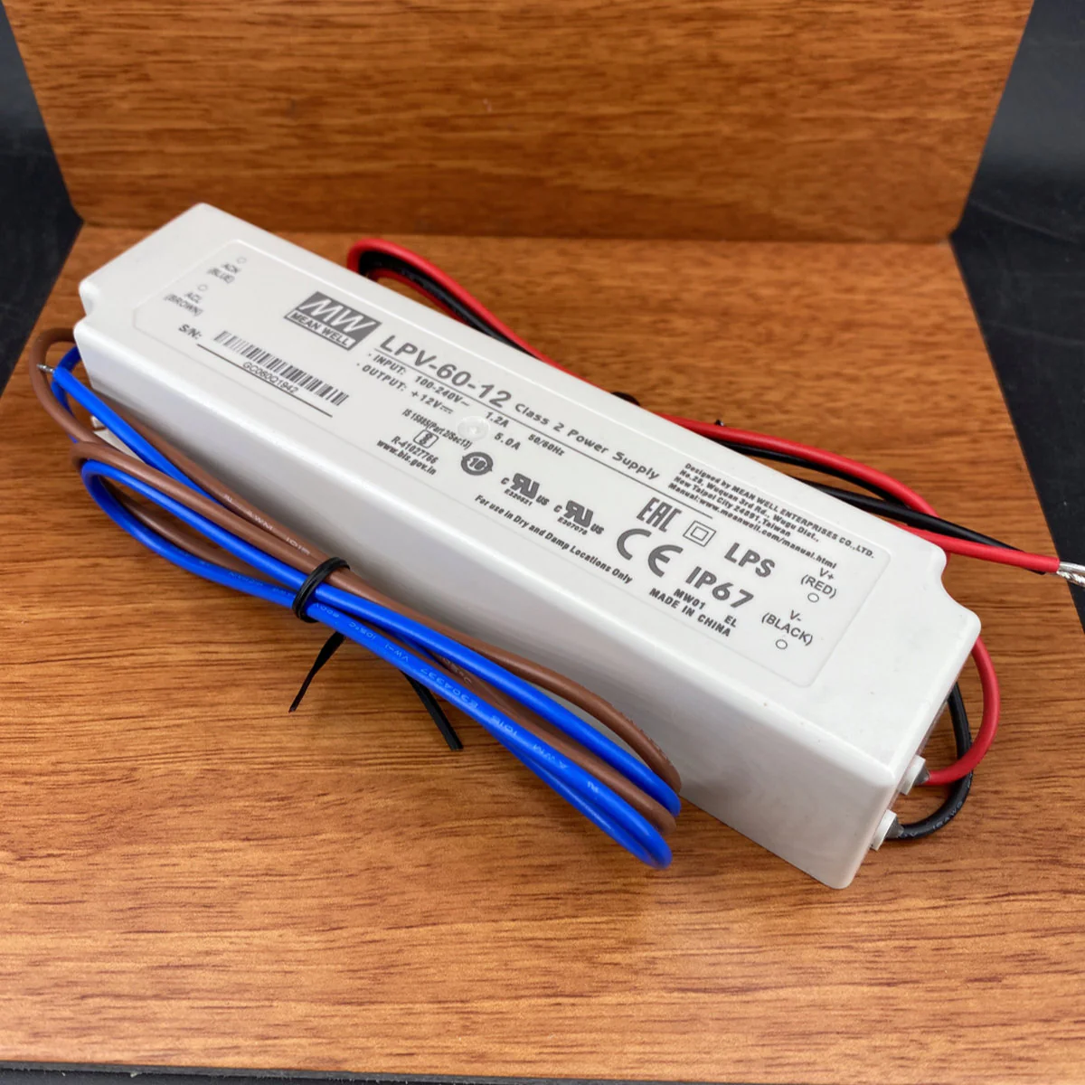 A Compact IP67 Waterproof LED Driver (5V) from the Meanwell LPV Series with attached red, black, and blue wires is shown on a wooden surface. Technical specs and certification labels are printed on its white casing for outdoor use.