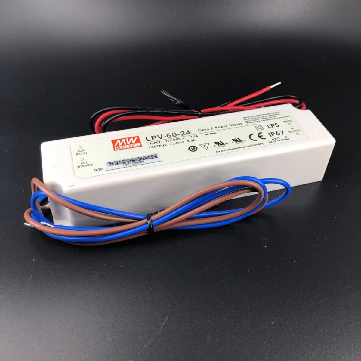 A white Compact IP67 Waterproof LED Driver (24V) ~ Meanwell LPV Series with red, black, blue, and brown wires rests on a black surface. Its label shows specs and safety icons. Suitable for outdoor use with attached wires for easy installation.