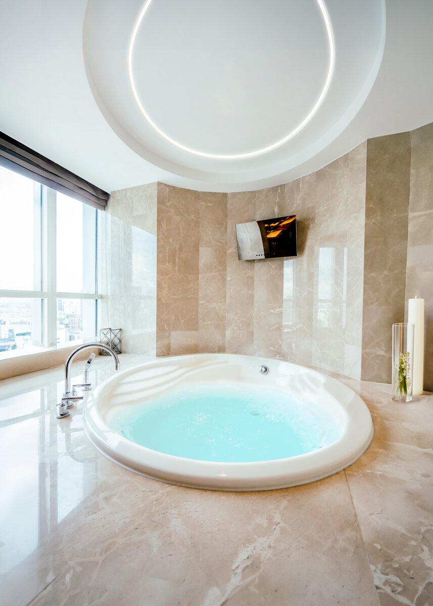A luxurious bathroom features a round built-in tub, beige marble walls and floors, large windows with city views, a wall-mounted TV, tall decorative candles, and recessed 1.05 Bendable Drywall LED Lighting Channel (Model Klus KOZUS-BD).
