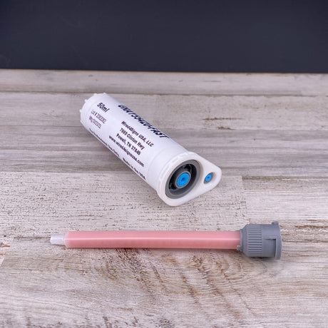 A Gray Fast-Cure 2-Part Methacrylate Adhesive ~ W4S 55420 (50ml 10:1 Mix) cartridge with printed text lies beside a pink mixing nozzle with a gray cap on light wood—ideal for aluminum bonding.
