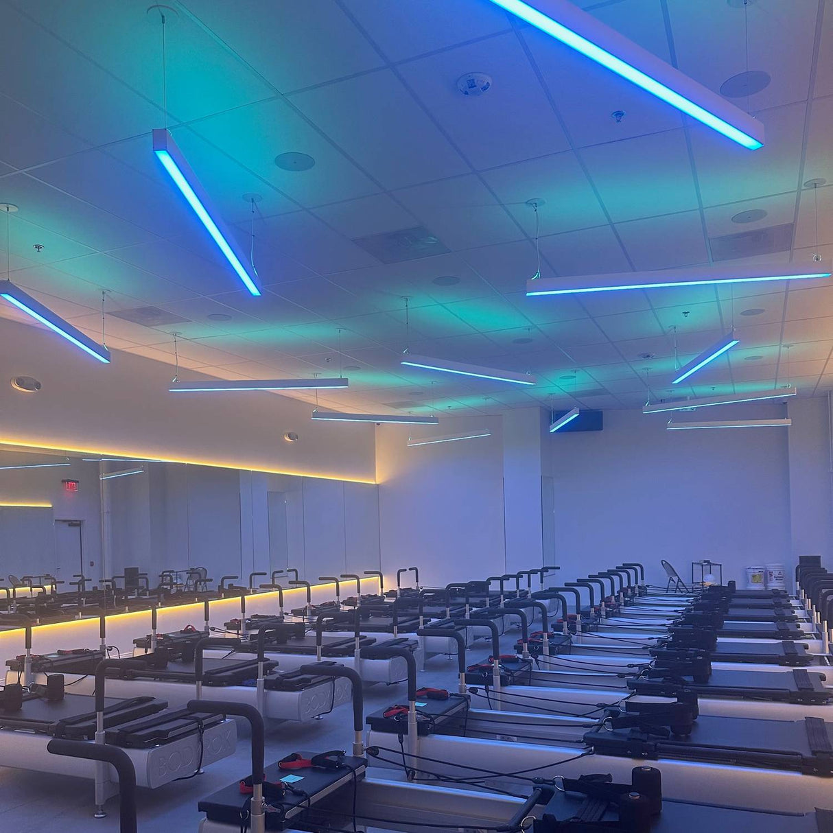 A modern fitness studio features rows of Pilates reformers under Munich Magnum 3.5x2.2 linear suspension lighting, a mirrored wall on the left, and a clean, minimal design with blue and green ceiling lights.