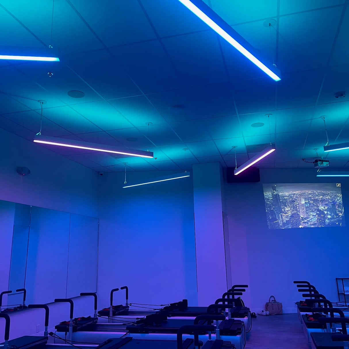 A fitness studio with reformer Pilates machines and blue-purple neon lights features the 3.5x2.2 Linear Suspension Lighting Channel ~ Model Munich Magnum on the ceiling, along with wall projections and mirrors reflecting the vibrant space.