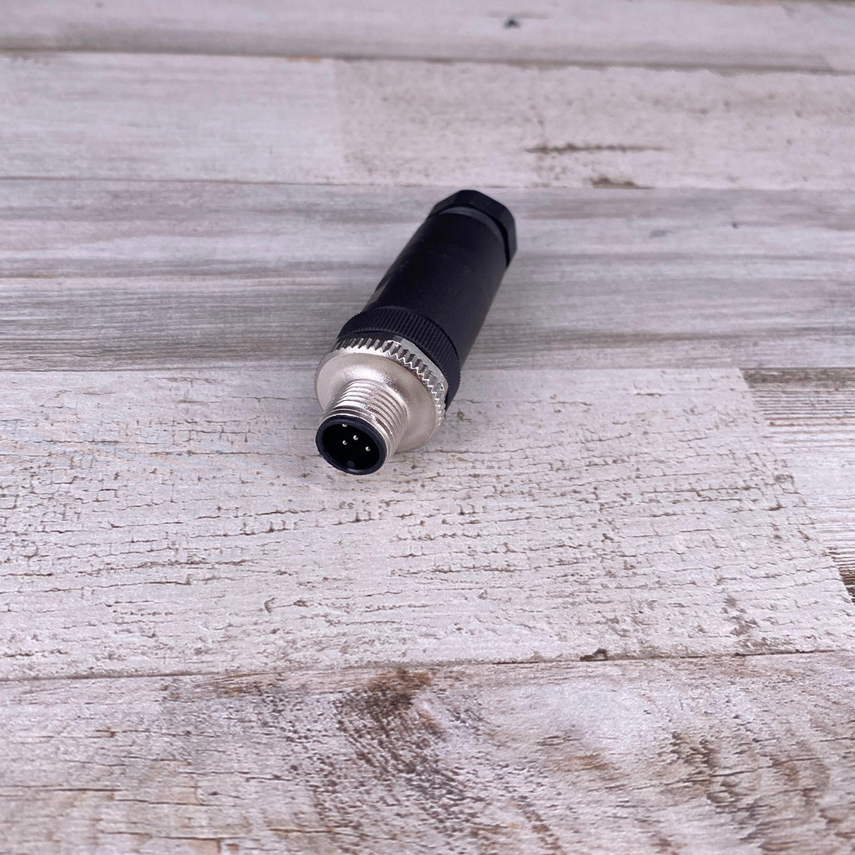 A close-up of the RGBW Male 5-pin Waterproof M12 Connector in black and silver, lying horizontally on a textured light wood surface with its pin end facing the camera. Commonly used for RGBW lighting setups.