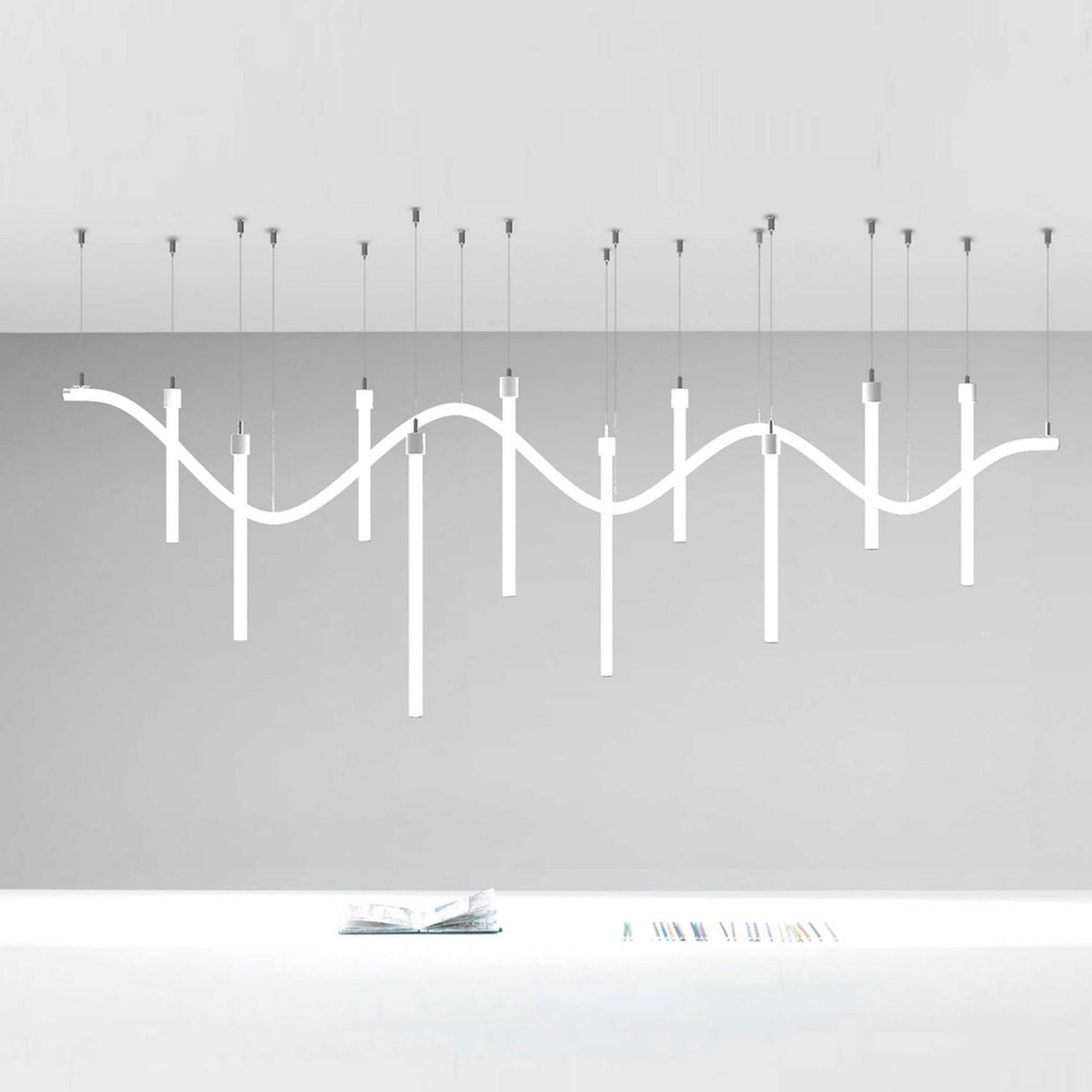 The Horizontal Hanger for Neon 360 Series is a modern white ceiling light with wavy lines and vertical bars, elegantly suspended by subtle cables above a white desk in a minimalist space.