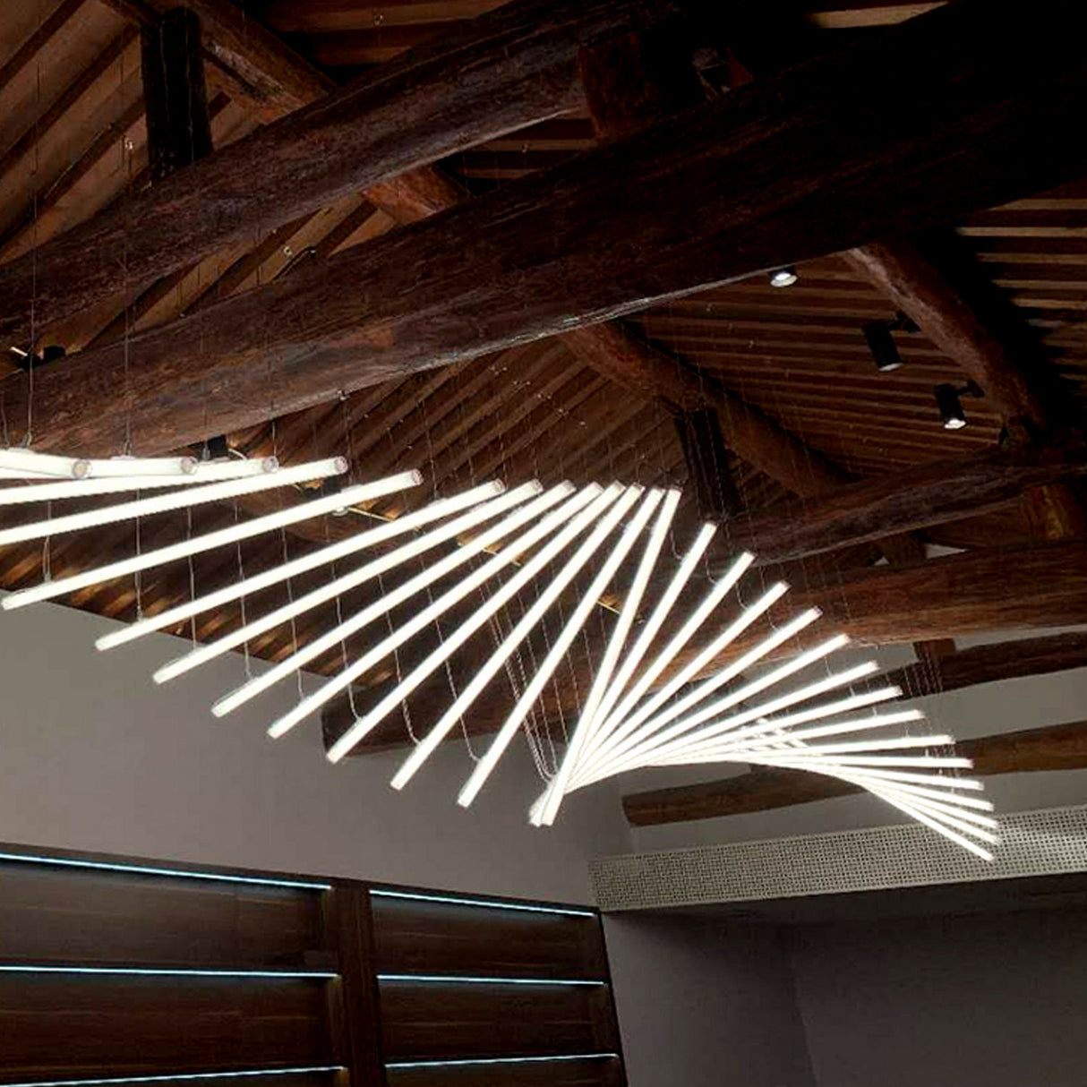 A Flexible White LED Neon Tube from the Neon 360 Series forms a spiral chandelier beneath exposed wooden beams, providing all-around illumination while natural light enters through horizontal windows on the left.