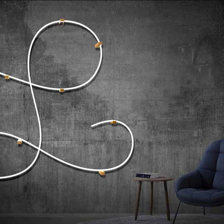A minimalist room with a dark concrete wall features the Flexible White LED Neon Tube ~ Neon 360 Series, providing 360-degree illumination, and a small round table with a book beside a blue upholstered chair.