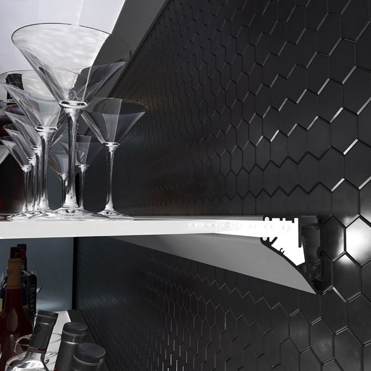 A Shelf Bracket Edge-Lit LED Channel (Model Pula P10) is mounted with martini glasses and bottles on a white shelf against a black hexagonal tile wall in a modern kitchen or bar setting.