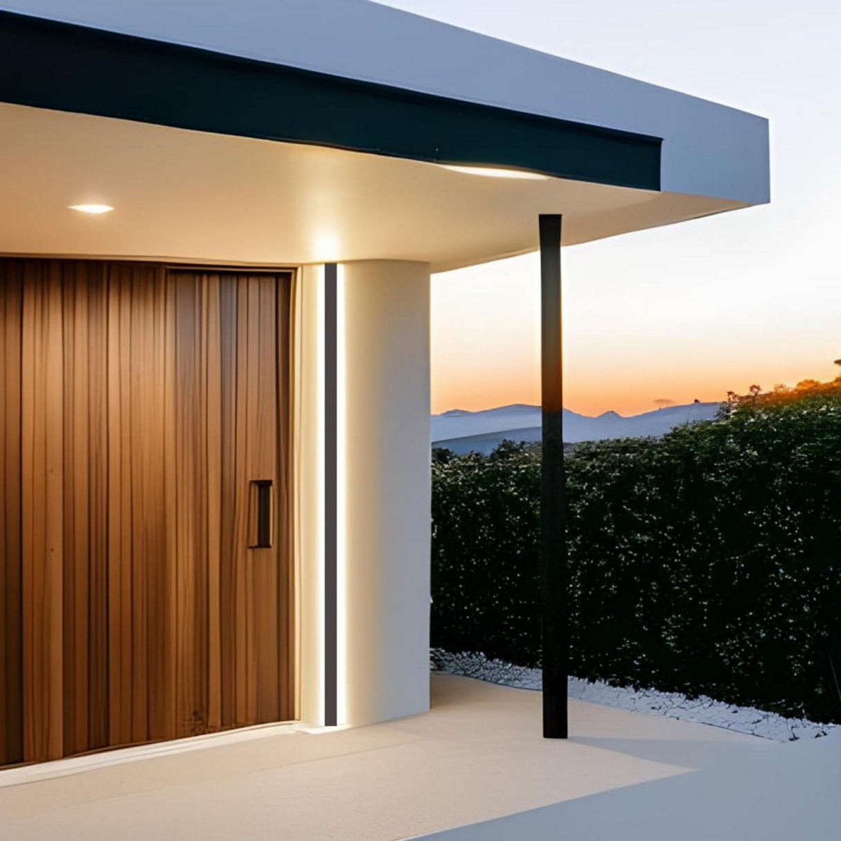 Modern house entryway at sunset with a large wooden door, sleek white walls, a thin black post, and elegant Direct/Indirect Linear Pendant Lighting LED Channel Model DPL70FL. Hedges in the background with distant mountains.