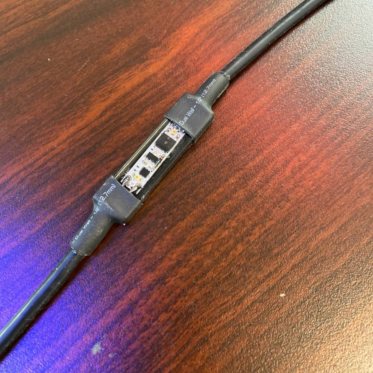 A close-up of an LED strip spliced into a black power cable with heat shrink tubing, resting on a reddish-brown wooden surface—perfect for pairing with the Miniature Daylight Switch for LED Channel ~ twilightSwitch by BleBox.