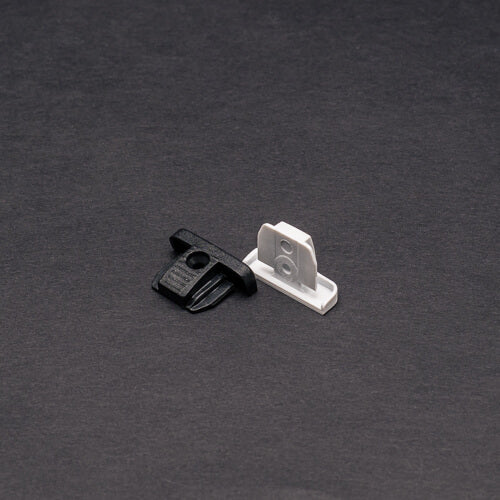 Two small plastic mounting clips, one black and one white, are shown on a dark surface. Designed for the End Cap for Power Track Twist, they have flat bases and upright tabs with center holes for secure attachment.