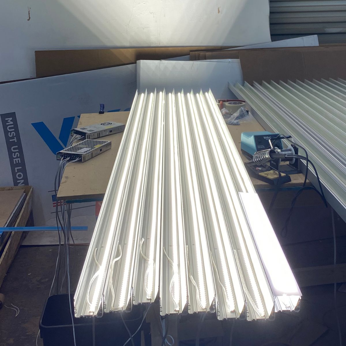 Several bright, white LED strips are arranged side by side in a Light Reflector 32mm Vega95 for DPL55-FL, DPL70-FL, PL55-FL, PLW70-FL, RPL55US, RPL70US and RPLW80FL on a workbench with tools and materials in the background.