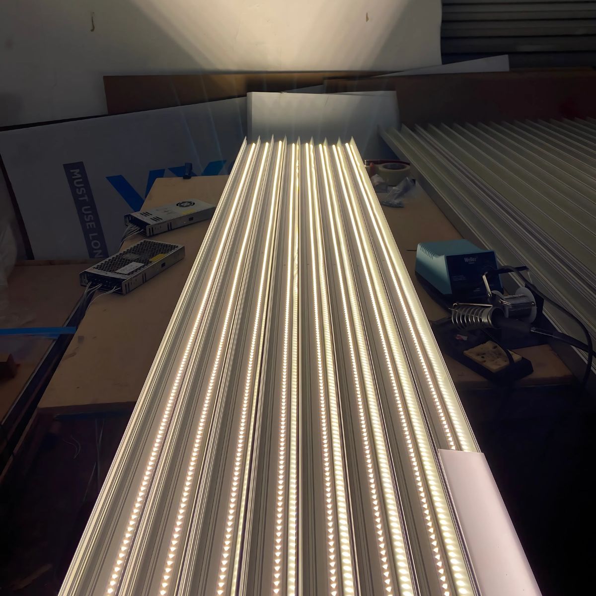 Rows of bright Light Reflector 32mm Vega95 units for DPL55-FL, DPL70-FL, PL55-FL, PLW70-FL, RPL55US, RPL70US, and RPLW80FL are neatly arranged on a workbench with tools and materials in the background. The lights cast a strong glow.