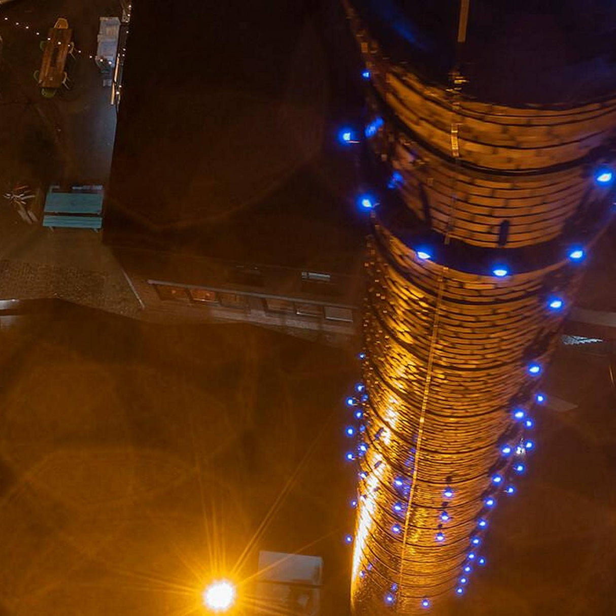 A tall brick tower glows orange-yellow, lit by a White Outdoor LED Wall Washer Light ~ Revo Basic Magno Dim Series with blue accents. The ground below remains dim, showing faint outlines of nearby structures and a streetlamp.