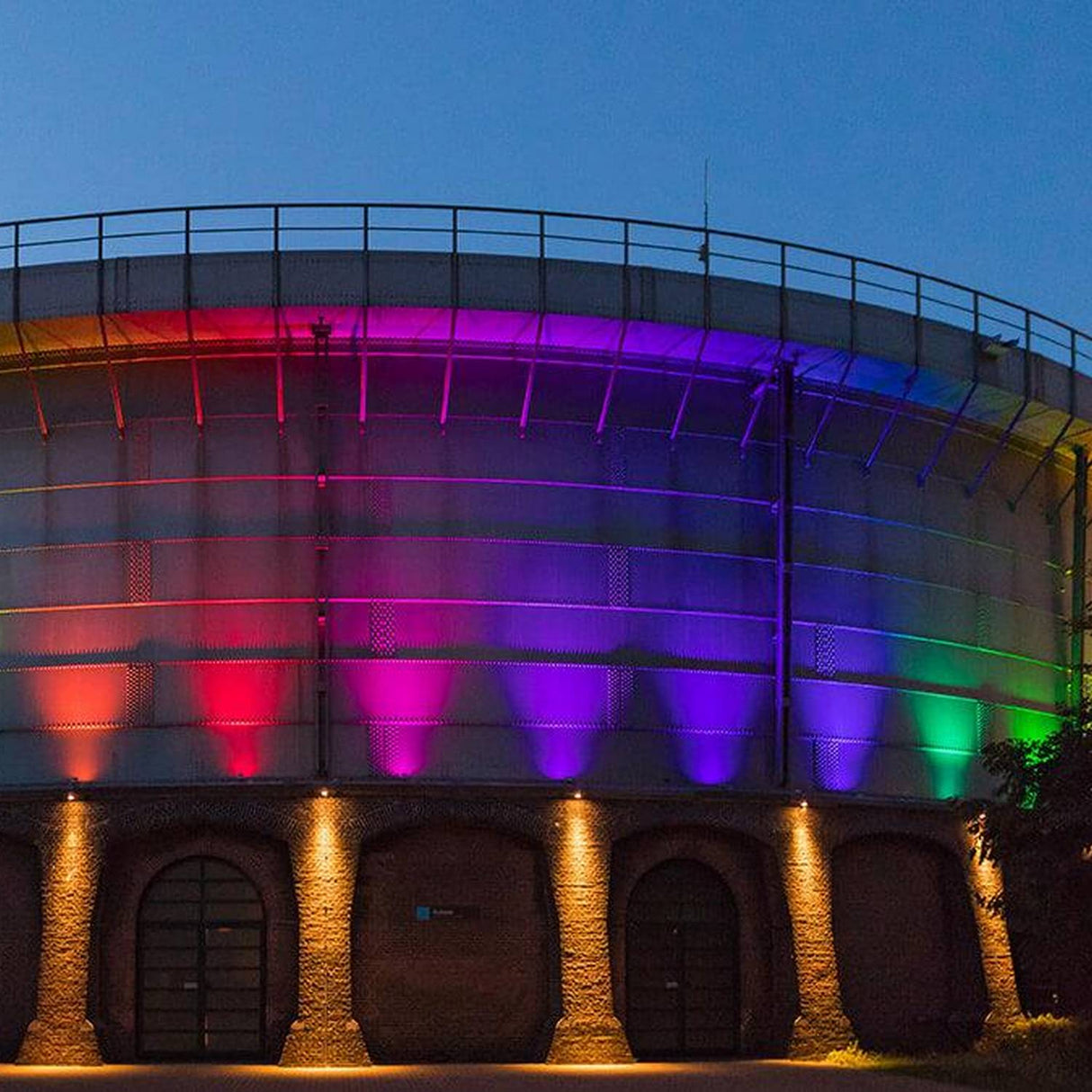 A large round brick building with arched doorways is illuminated at dusk by Color-Changing Outdoor LED Wall Washer Lights (Revo ColorFlow Casambi Series), casting vibrant rainbow hues against the evening sky.