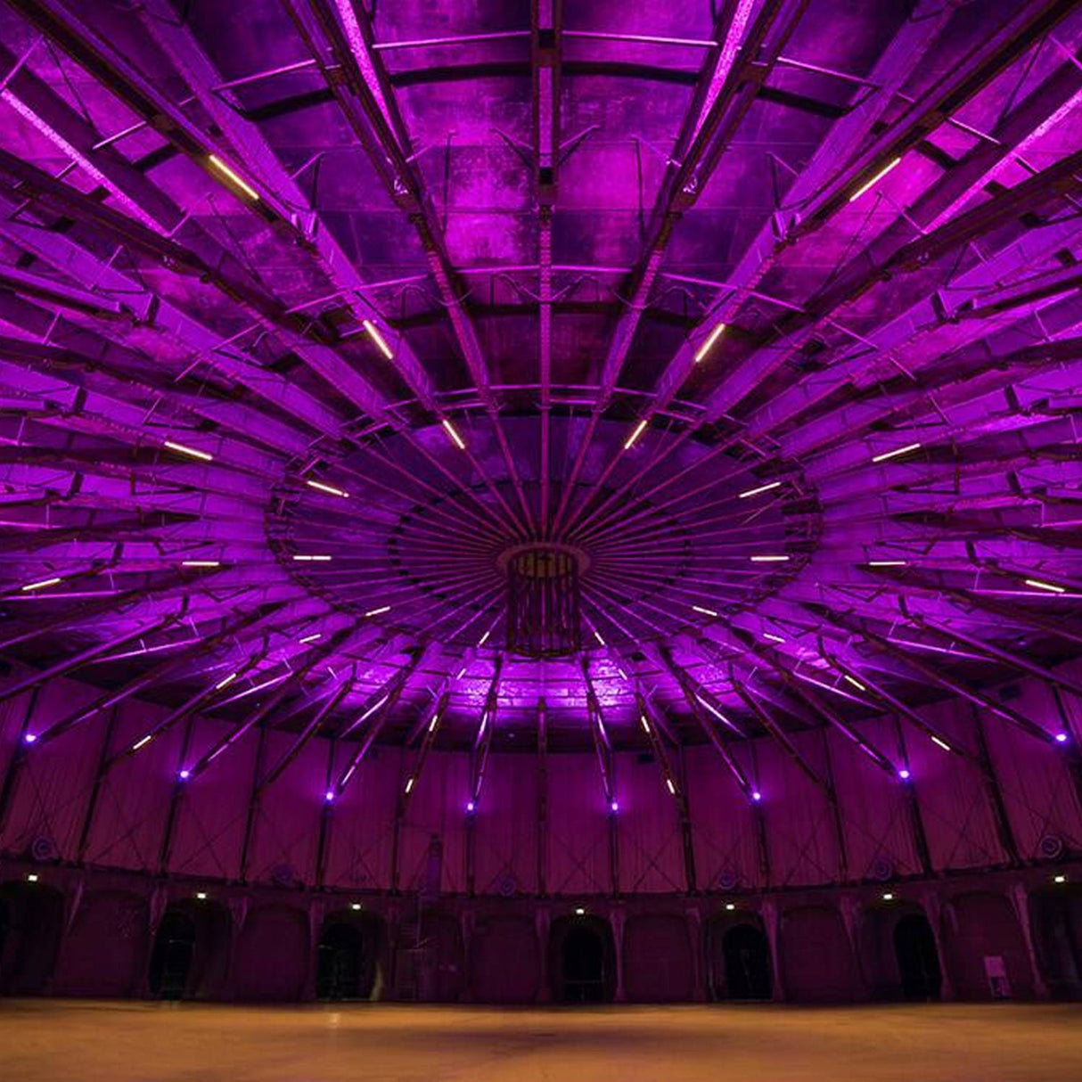 A spacious, circular indoor venue with a high dome ceiling is dramatically illuminated by vibrant purple from the Revo ColorFlow Casambi Series Color-Changing Outdoor LED Wall Washer Light, highlighting the structure’s industrial symmetry.