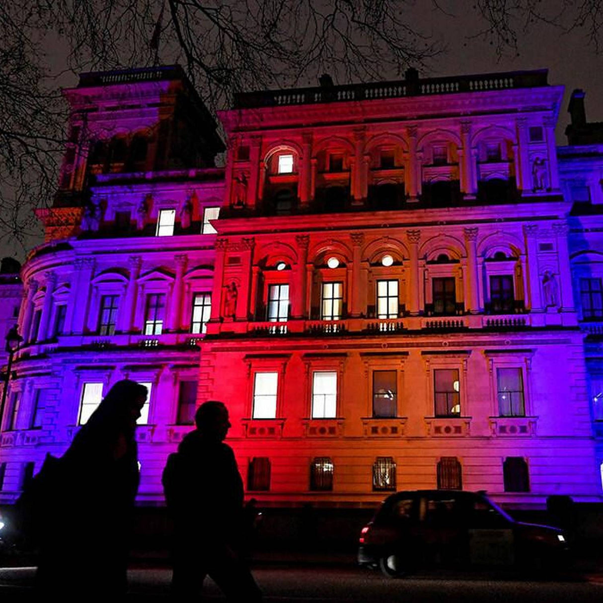 At night, a large building is illuminated in blue, purple, and red by the Color-Changing Outdoor LED Wall Washer Light ~ Revo ColorFlow Casambi Series. Two people and a car are silhouetted in front as bare branches hang overhead.