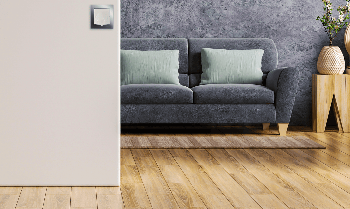 Modern living room with a grey sofa, two light green pillows, a wooden side table, and a gray textured wall. The Cable Outlet With Kontak Simon Frame ~ cableoutlet by Blebox is near the light switch on the light wooden floor.