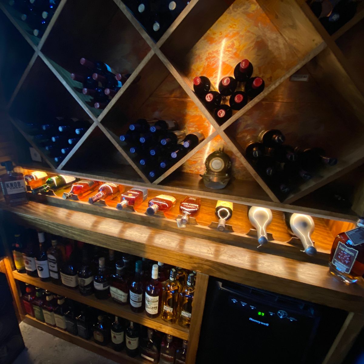 A dimly lit wine cellar features crisscross wooden shelves with wine bottles and a counter displaying liquor beneath energy-efficient Slim LED Channel ~ Model Slim8 lighting, with more spirits below next to a small black fridge.