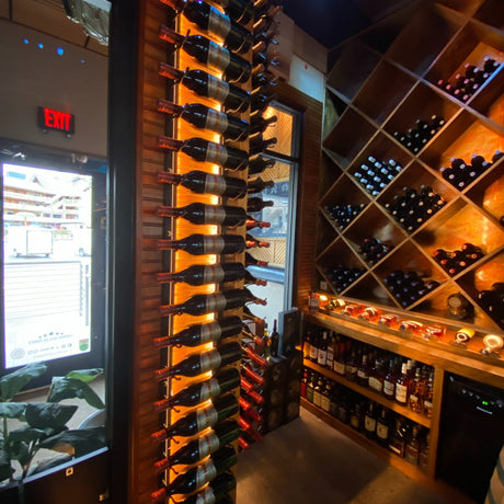 A wine cellar features racks of wine bottles and shelves of liquor, illuminated by energy-efficient Slim LED Channel ~ Model Slim8 lighting, next to a glass door with daylight outside.