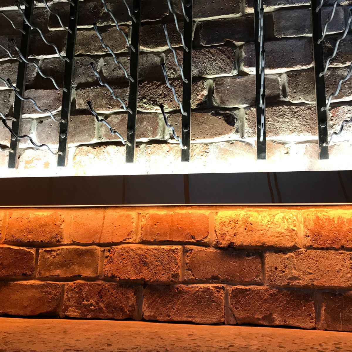 Brick wall with metal wine racks and horizontal shelves, featuring a surface-mount Slim LED Channel ~ Model Slim8—one shelf shines white light, the lower glows warm orange, creating an energy-efficient contrast on textured brick.