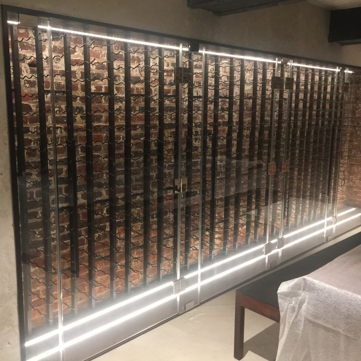 A modern wine cellar with glass doors, metal racks, and brick walls is illuminated by energy-efficient white LED strip lights in a Slim LED Channel ~ Model Slim8, with a covered table in the foreground.
