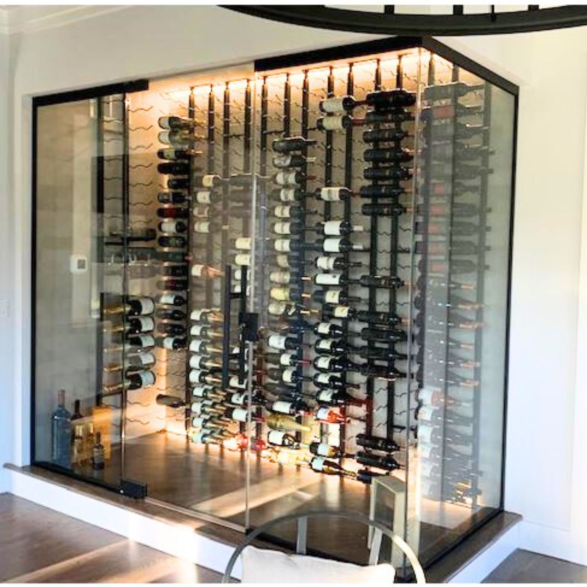 A modern glass-enclosed wine cellar features energy-efficient Slim LED Channel ~ Model Slim8 backlighting, multiple racks for different bottles, and large bottles showcased at the bottom—all integrated into a white wall.