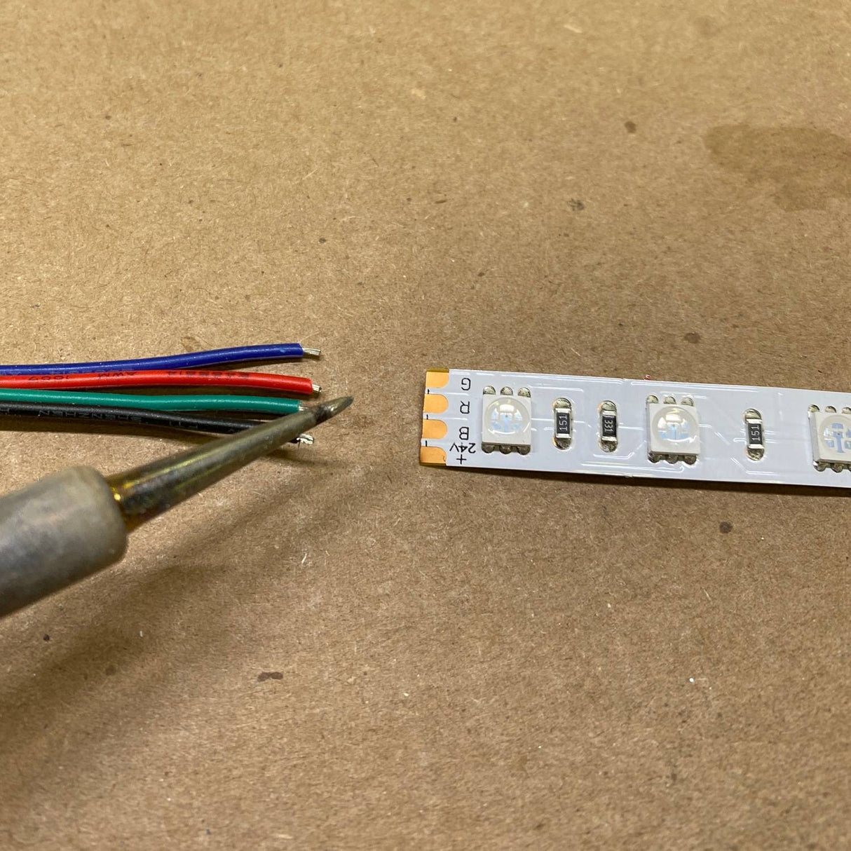 The Solder Joint - 4 conductors is shown with a soldering iron near four colored wires (black, blue, green, and red) and LED strip connector pads on a brown surface, ready for precise soldering.