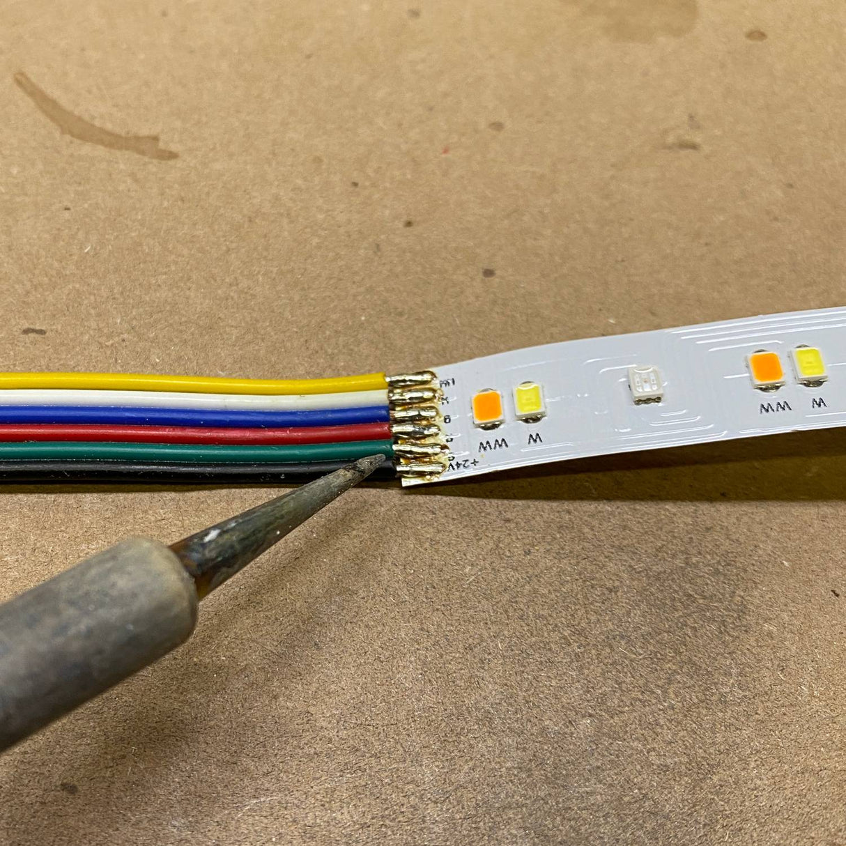 A close-up of a Solder Joint - 6 conductors, showing a soldering iron forming a clean connection on multicolored wires attached to an LED strip on a brown surface.