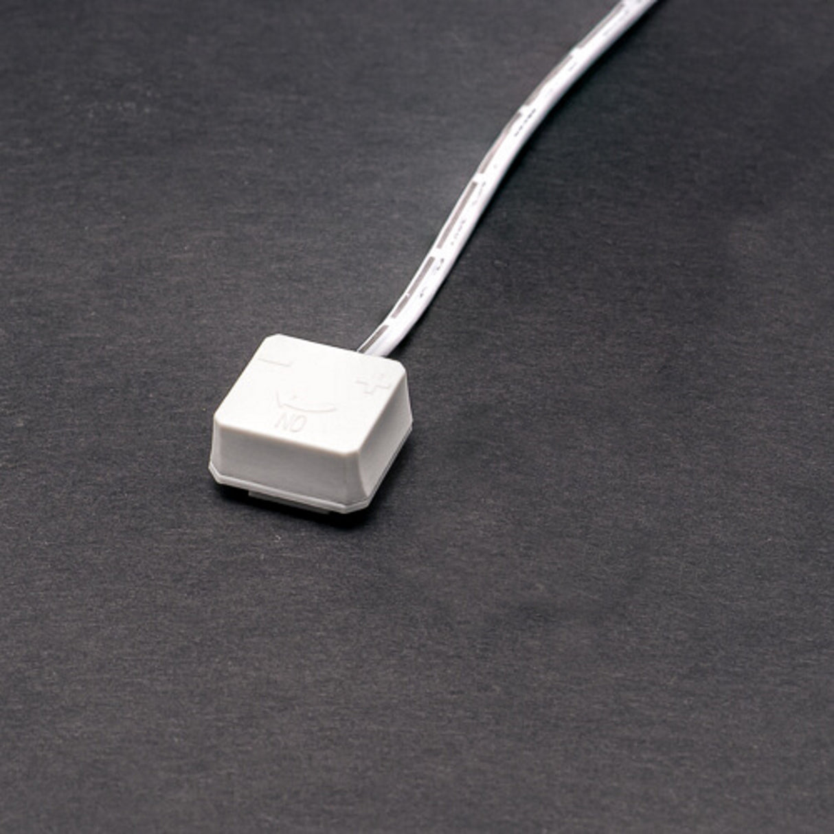 A Power Track Twist Plug Connector, a white switch with a short wire attached, rests on a dark gray surface. Designed for LED fixtures, it features an on/off symbol and plus/minus signs on its top.