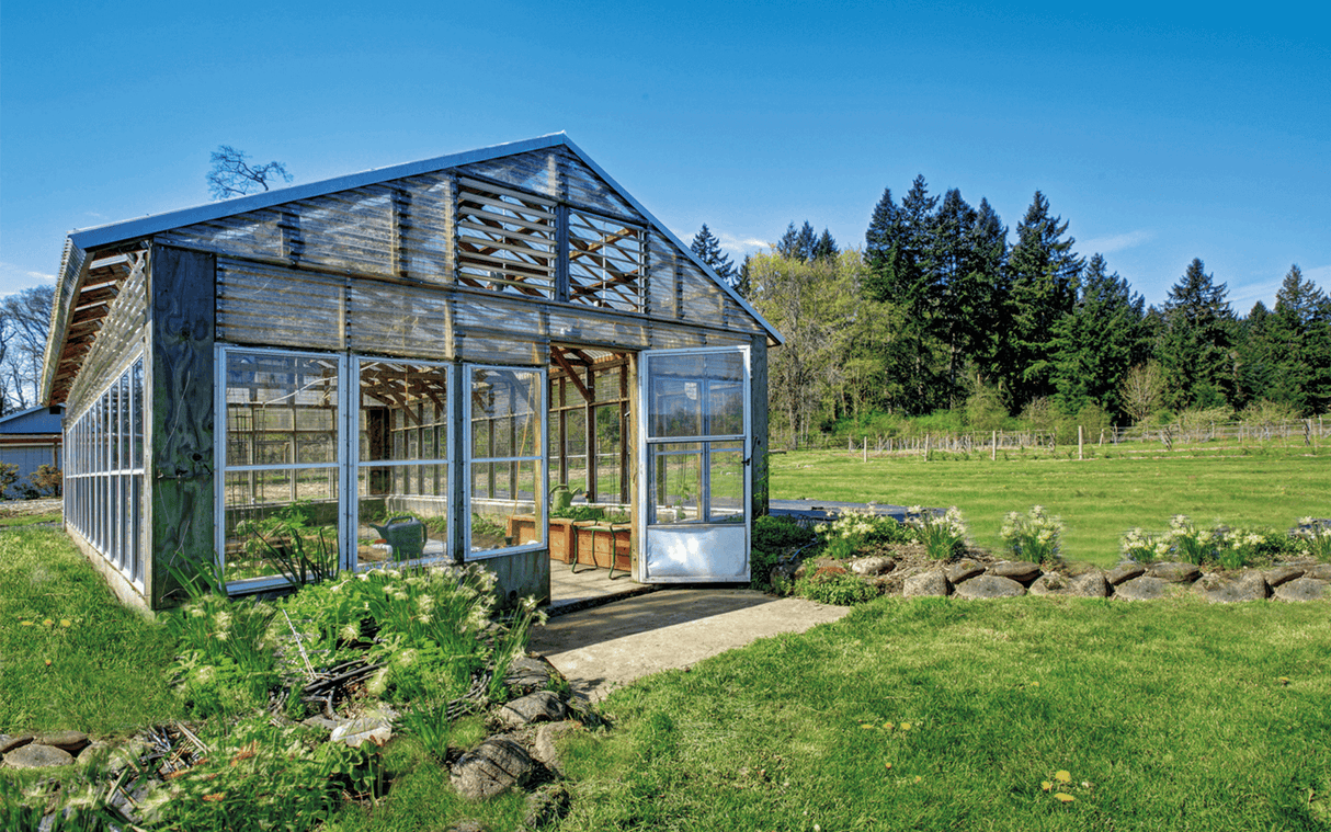 A weathered glass-walled greenhouse stands on a grassy lawn among rocks and plants, using the tempSensor by Blebox smart wireless temperature sensor for real-time monitoring, set against trees and a clear blue sky.