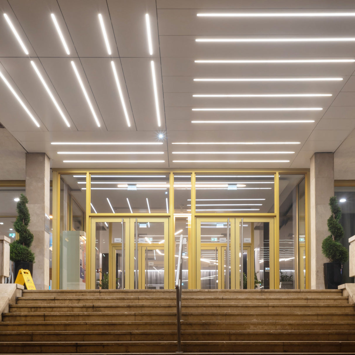 A wide staircase leads to glass doors framed in yellow. Above, the ceiling features modern linear lighting using the 1.3” Deep-Section Recessed LED Channel (Model Vario30-07), while potted topiary plants flank the stylish entrance.