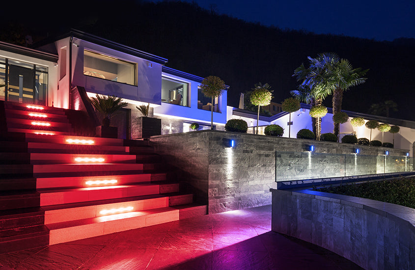 A modern house exterior at night features illuminated red and white stair lights controlled by the Outdoor RGBW 4-Ch Wi-Fi LED Controller ~ wLightBox Pro by BleBox, glass railings, palms, and trimmed bushes for a vibrant, inviting look.