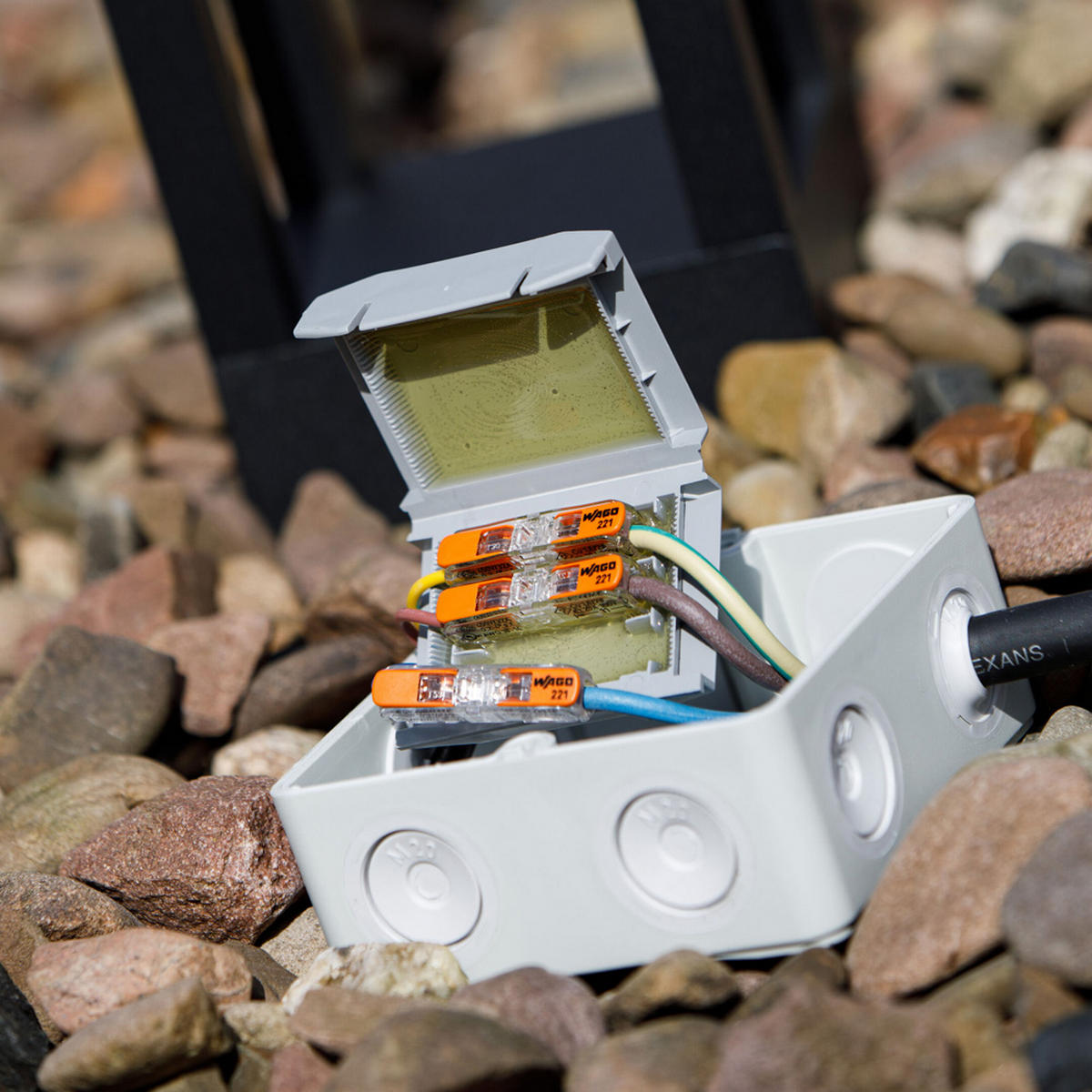A Waterproof Sealing Gelbox for Wago 221 Inline Lever-Nut Connectors sits open on rocks, displaying orange wire connectors and several connected wires inside.