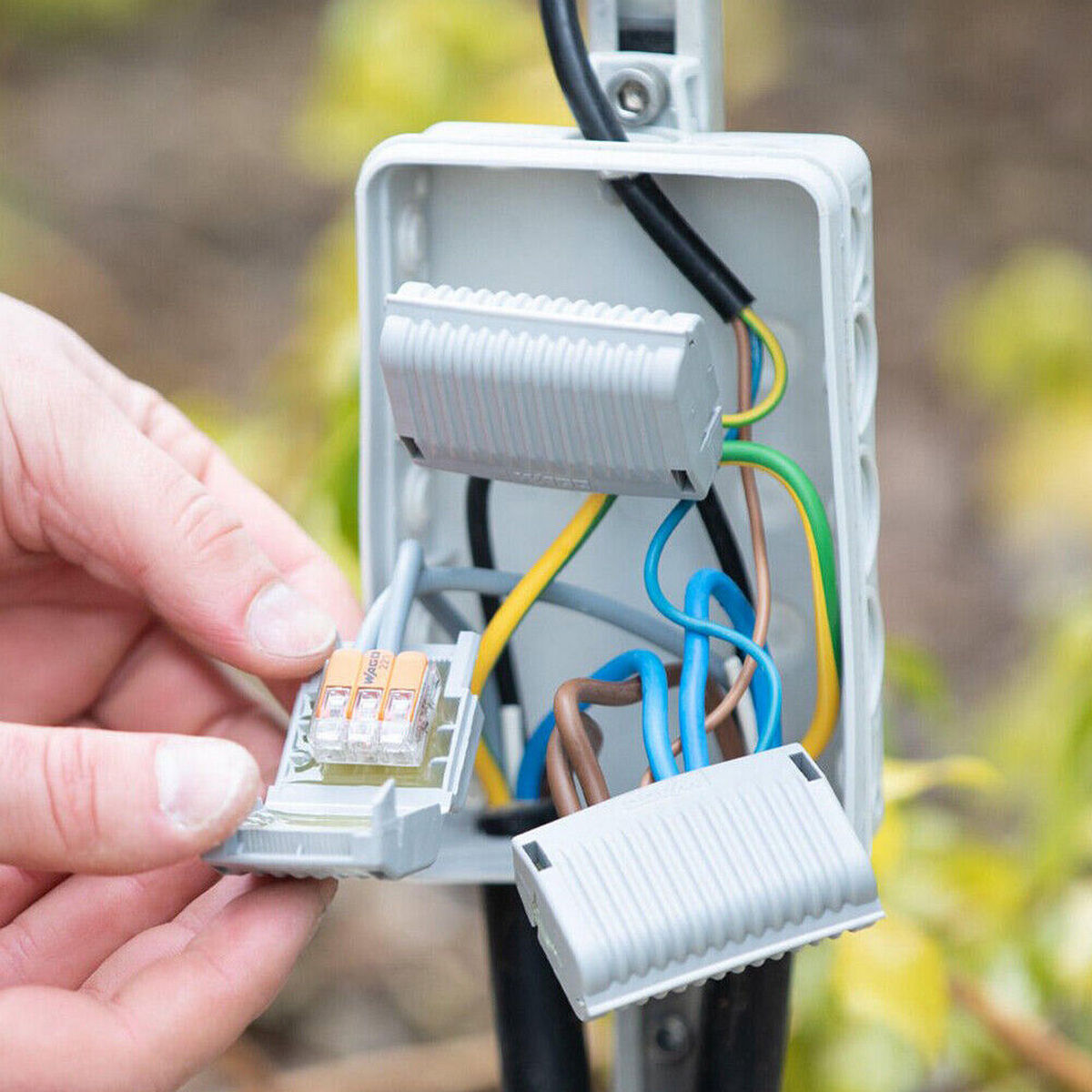 A person connects colored wires to a terminal block inside an open Waterproof Sealing Gelbox for Wago 221 Splicing Lever-Nut Connectors, ensuring moisture protection. Green plants are visible blurred in the background.