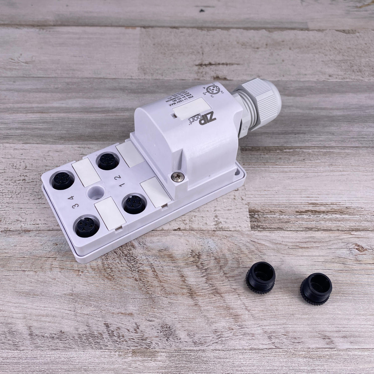 The Waterproof Junction Block for RGBW LED Lights (ZipPort JBH Series) is a white plastic box with IP67 protection, six numbered black cable entry points, a cable gland, and two loose black caps on a light wood surface.