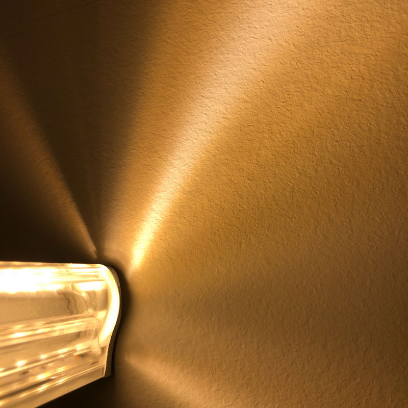 A close-up of the LED Strip Vaulted Ceiling Light Channel (Model Alu-Asymmetric), wall-mounted and casting warm yellow, fan-shaped rays and shadows across a textured beige wall with its unique asymmetric LED profile.