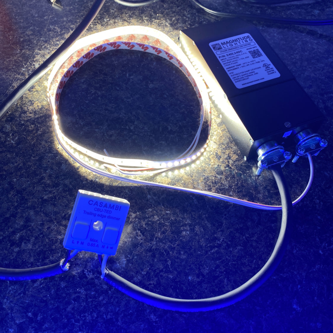 A coiled LED light strip glows on a dark countertop, powered by a black supply unit and the Casambi Bluetooth Controller Model CBU-TED-LR for seamless wireless lighting control via attached wires.