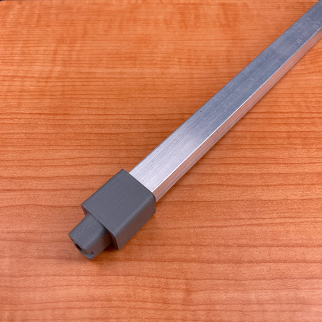 An Aluminum square tube for light sticks, 1/2 inch wide, with a gray rectangular plastic cap on one end, is placed diagonally on a light brown wood-grain surface.