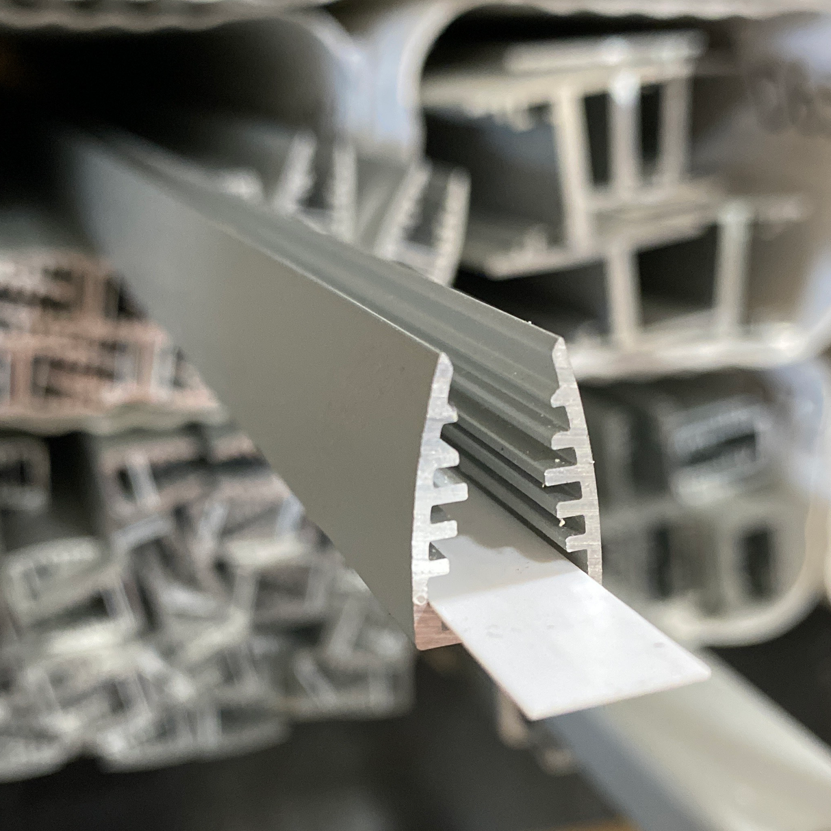 Close-up of a stack of aluminum profiles, with a grooved, rounded edge in front—perfect for the 1/4 LED Edge-lit Acrylic Sign Base Channel (Model Mikro10)—while other metal profiles remain blurred in the background.