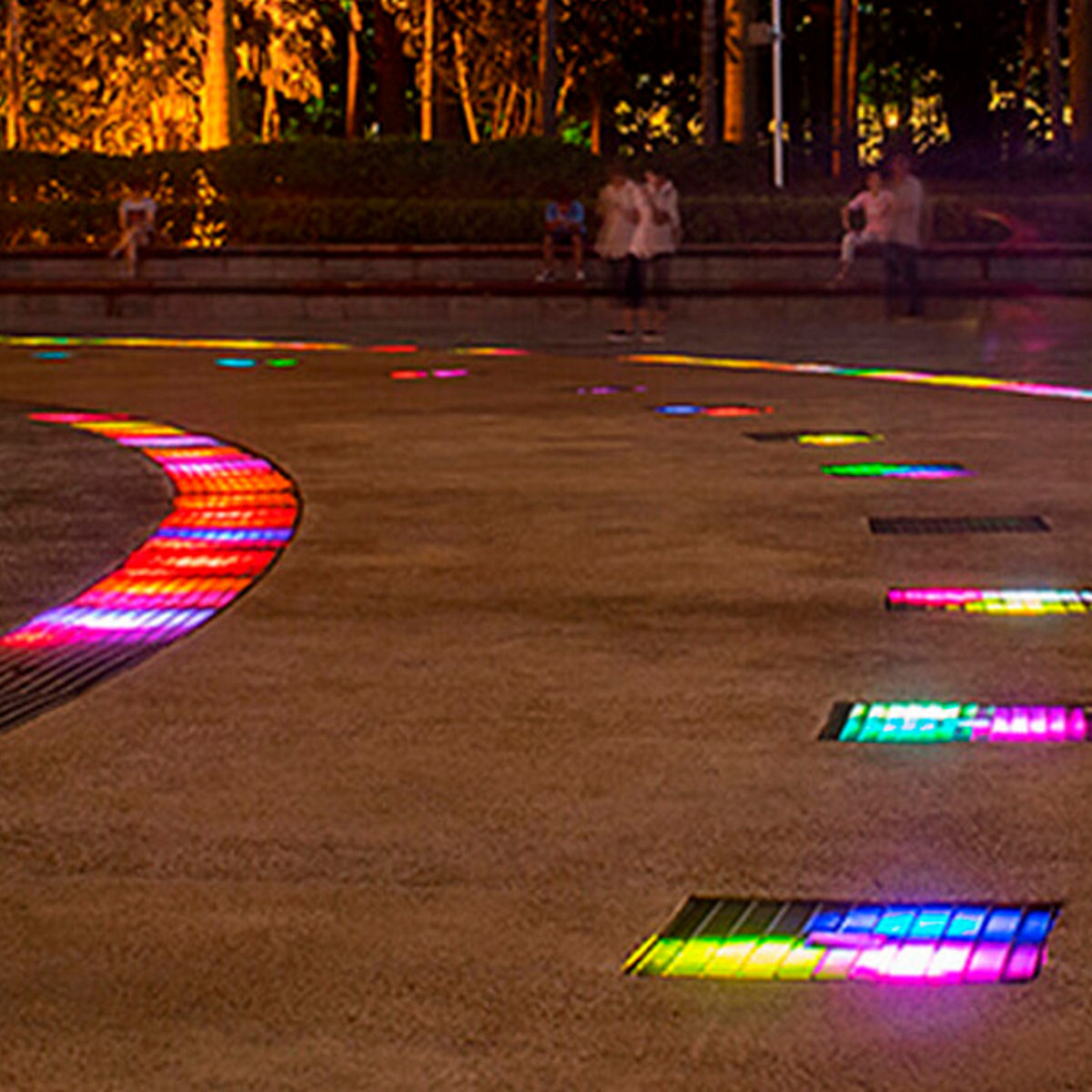 At night in a public square, vibrant illuminated tiles embedded in the ground mimic the patterns of the Blebox Garage Parking Assist and Occupancy Sensor. People are seated or standing nearby amidst trees and benches.