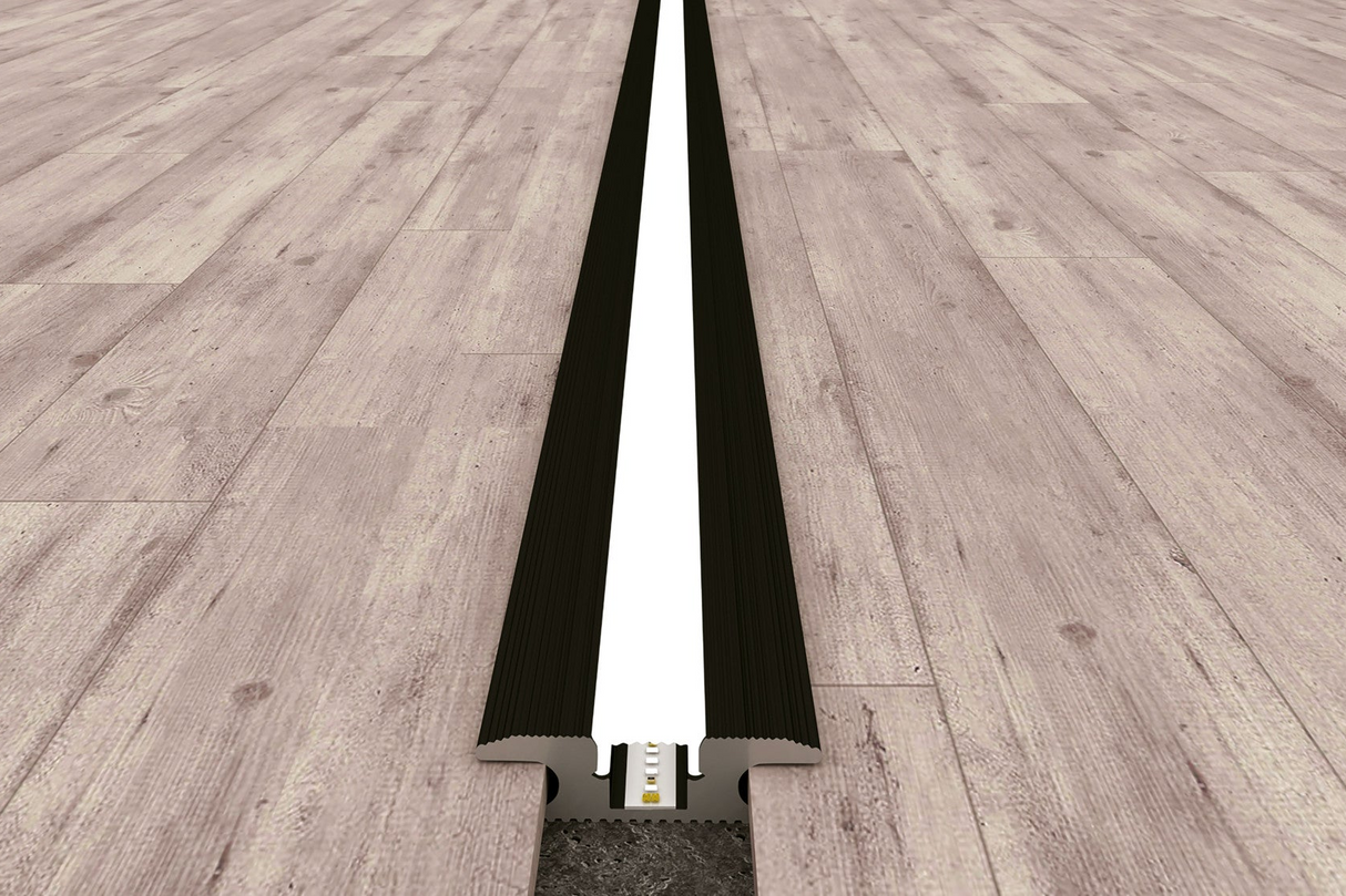 A close-up of the Floor Light Profile ~ Model Estambul, a treadable LED channel with black and white trim, seamlessly integrated between light wood planks for a modern look.