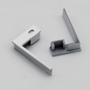 Two L-shaped white End Caps for Tallin Profile lighting are displayed on a light gray surface—one with a hole and the other featuring grooved end caps to enable versatile mounting.
