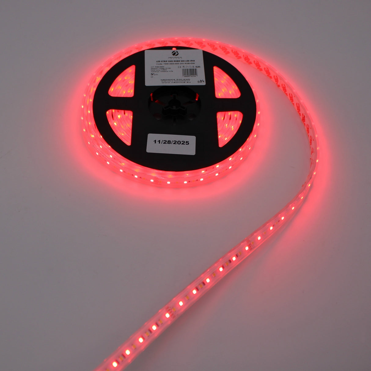 A partially unrolled spool of Low-Power RGBW IP67 Waterproof LED Strip (24V) ~ Iris Series glows against a plain light background, with the reel labeled 11/28/2025. The energy-efficient lights shine brightly.