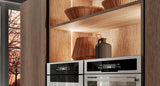 Wooden kitchen shelves with decorative bowls, a vase, and built-in ovens are illuminated by the Recessed Angular LED Channel ~ Model Amsterdam Mini. Warm lighting highlights the shelves, with a sunset-view window on the left.