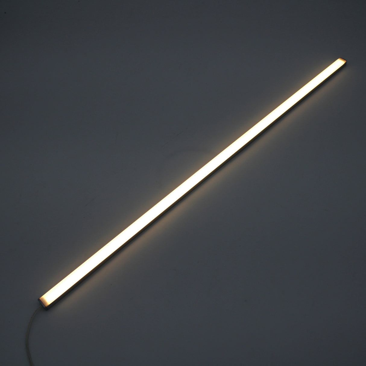 The White Universal Surface-mount LED Lighting Fixture ~ Begton12 Series, a long, slim fixture, emits a bright, dotless white glow and is placed diagonally against a dark background.