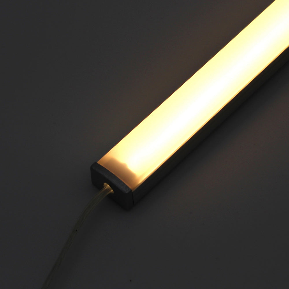 A close-up of the White Universal Surface-mount LED Lighting Fixture ~ Begton12 Series glowing warmly as under-cabinet lighting, with a power cord attached at one end, set against a dark background.