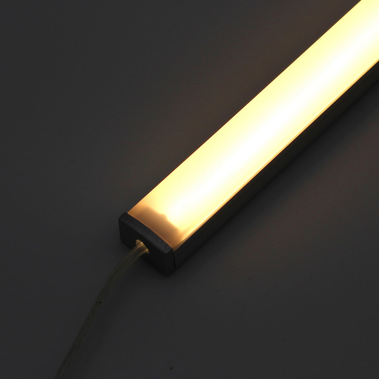 A close-up of the White Universal Surface-mount LED Lighting Fixture ~ Begton12 Series glowing warmly as under-cabinet lighting, with a power cord attached at one end, set against a dark background.