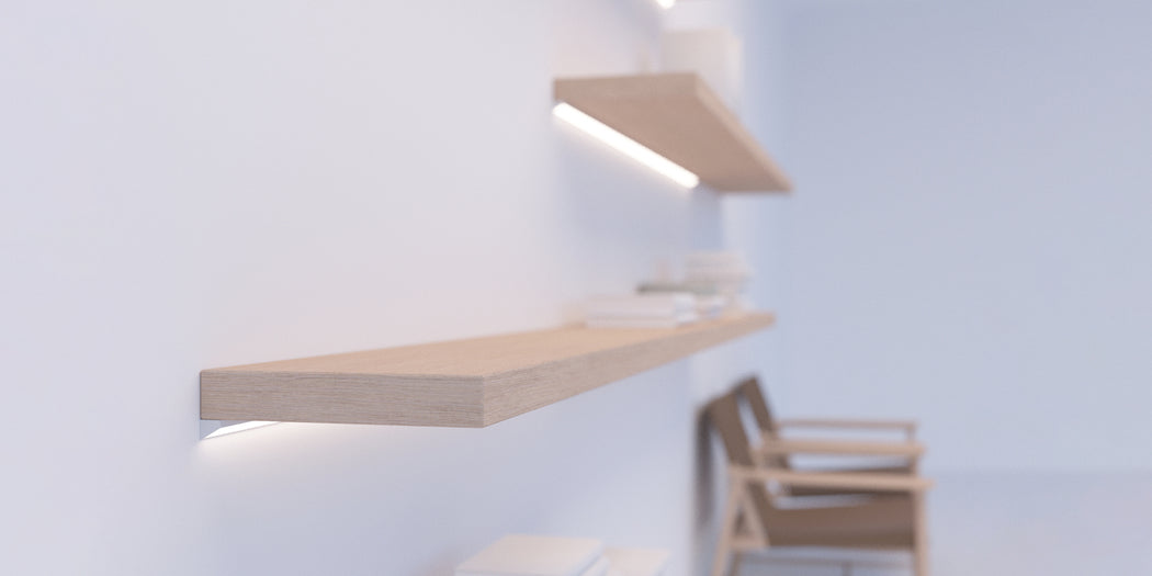 A minimalist interior showcases light wood floating shelves mounted on a white wall, with books neatly arranged and a wooden armchair in the background. The 30/60 Degree Corner LED Channel ~ Model Corner10.v2 runs subtly along the shelving, providing modern accents and gentle illumination. The overall atmosphere is soft and bright.