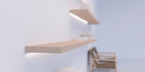 A minimalist interior showcases light wood floating shelves mounted on a white wall, with books neatly arranged and a wooden armchair in the background. The 30/60 Degree Corner LED Channel ~ Model Corner10.v2 runs subtly along the shelving, providing modern accents and gentle illumination. The overall atmosphere is soft and bright.