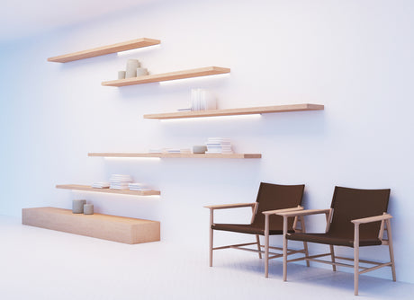 A minimalist room features floating wooden shelves on a white wall, illuminated by Surface Mount Corner LED Channel ~ Model Corner12.v2. Nearby, two modern black chairs with wooden armrests add a stylish and cozy touch.